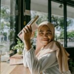 A smiling woman in a hijab reading a book by a window in a cozy cafe in Banda Aceh, Indonesia.