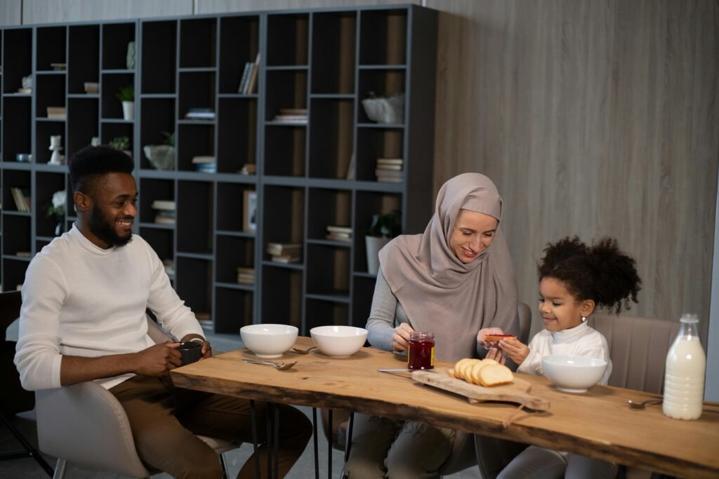 Positive African American father drinking hot beverage while Muslim mother in hijab giving sandwich with jam to black daughter sitting at table and having breakfast